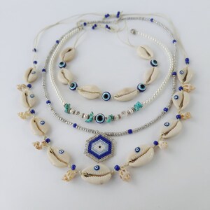 May include: Layered necklace with white seashells, blue evil eye beads, and a silver chain. The necklace also features a hexagonal charm with a blue and white evil eye design.