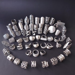 May include: A collection of silver-toned rings with various designs, including geometric shapes, floral patterns, and abstract forms. The rings are arranged in a circular pattern on a black background.