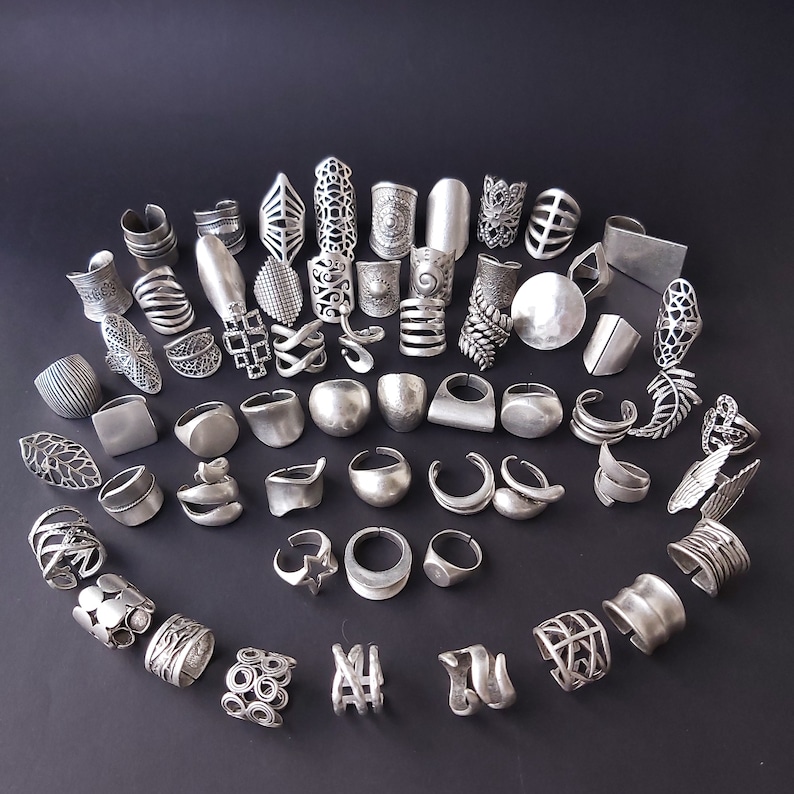 May include: A collection of silver-toned rings with various designs, including geometric shapes, floral patterns, and abstract forms. The rings are arranged in a circular pattern on a black background.