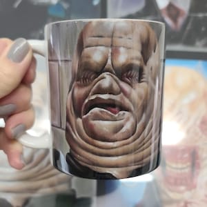 May include: White ceramic mug with a graphic of a creature with a large head and a wrinkled face. The creature has a large mouth and a tongue sticking out.