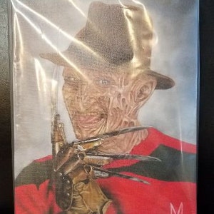 May include: A close-up of Freddy Krueger, the iconic horror character, wearing a red and black striped sweater and a brown fedora. He is holding a glove with razor-sharp blades. The image is in a plastic sleeve.