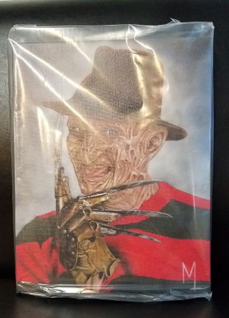 May include: A close-up of Freddy Krueger, the iconic horror character, wearing a red and black striped sweater and a brown fedora. He is holding a glove with razor-sharp blades. The image is in a plastic sleeve.