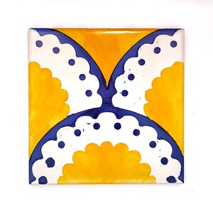 May include: Square ceramic tile with a hand-painted design. The tile features a symmetrical pattern with a yellow background and a butterfly-like design in white and blue. The white sections have a scalloped edge and blue dots.