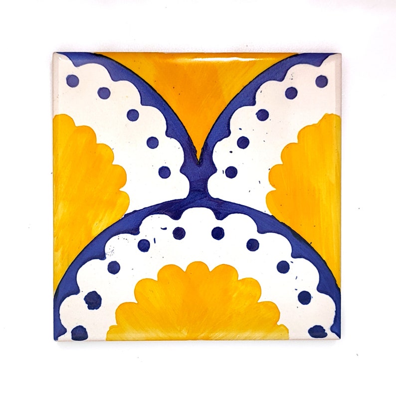 May include: Square ceramic tile with a hand-painted design. The tile features a symmetrical pattern with a yellow background and a butterfly-like design in white and blue. The white sections have a scalloped edge and blue dots.