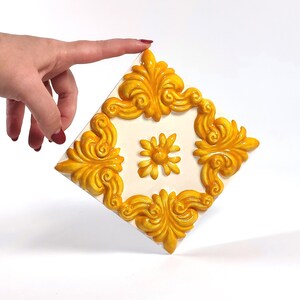 May include: A square, off-white decorative tile with a raised, ornate gold design. The gold design features a floral motif in the center and a repeating scroll pattern around the edges. The tile is held by a hand.