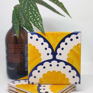 May include: Three square ceramic tiles with a yellow and white floral design, edged with navy blue. The tiles are stacked on a white surface, with a brown bottle and green plant in the background.