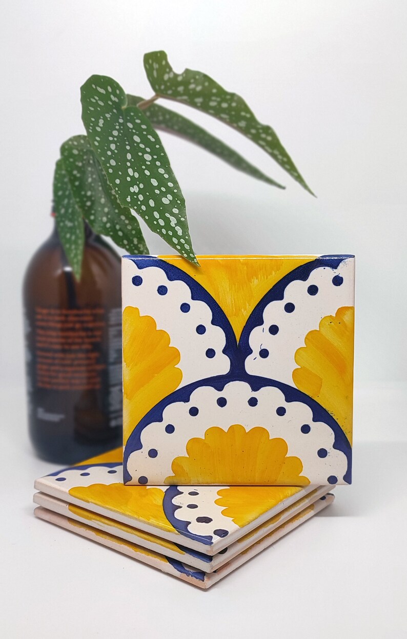 May include: Three square ceramic tiles with a yellow and white floral design, edged with navy blue. The tiles are stacked on a white surface, with a brown bottle and green plant in the background.
