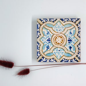 Puede incluir: Azulejo cerámico cuadrado con un diseño simétrico detallado en tonos azules, dorados y blancos. El azulejo presenta un motivo floral central y patrones de borde ornamentados. El azulejo mide unos 10 cm cuadrados.