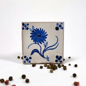 Puede incluir: Azulejo cerámico cuadrado con un diseño floral azul pintado a mano. La flor está centrada en el azulejo, con un tallo y hojas. Las esquinas tienen pequeños detalles decorativos azules. El azulejo está rodeado de granos de pimienta.