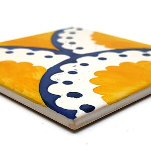 May include: Square ceramic tile with a hand-painted design. The tile features a yellow and white pattern with blue accents. The design is symmetrical, with four yellow sections separated by white and blue curved lines. The tile is viewed at an angle.