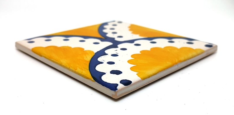 May include: Square ceramic tile with a hand-painted design. The tile features a yellow and white pattern with blue accents. The design is symmetrical, with four yellow sections separated by white and blue curved lines. The tile is viewed at an angle.