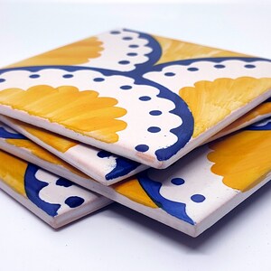 May include: Four square ceramic coasters stacked at an angle. Each coaster has a white base with a yellow and blue floral design. The coasters are hand-painted, showcasing a detailed pattern. The coasters are suitable for drinks.