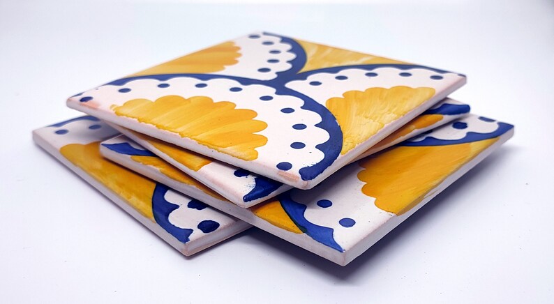 May include: Four square ceramic coasters stacked at an angle. Each coaster has a white base with a yellow and blue floral design. The coasters are hand-painted, showcasing a detailed pattern. The coasters are suitable for drinks.