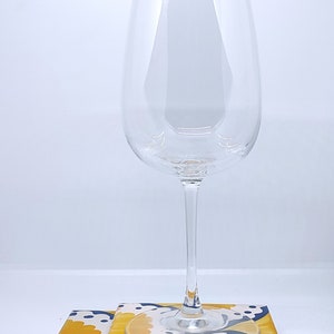 May include: A clear wine glass sits on a stack of square coasters. The coasters have a yellow and white floral design with blue accents. The wine glass is tall and slender. The coasters are approximately 10 cm square.