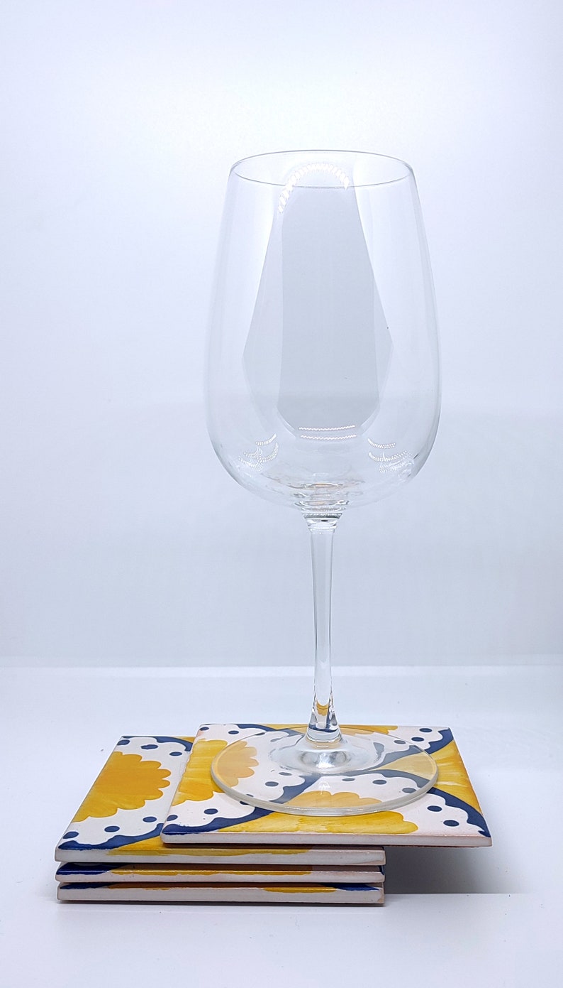 May include: A clear wine glass sits on a stack of square coasters. The coasters have a yellow and white floral design with blue accents. The wine glass is tall and slender. The coasters are approximately 10 cm square.