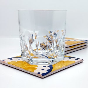 May include: A clear, cut-glass tumbler rests on a square coaster with a floral design. The coaster has a cream background with yellow and navy blue floral patterns. More coasters are stacked behind, indicating a set.