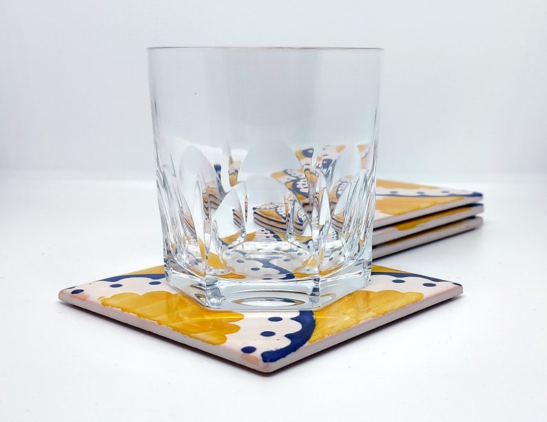 May include: A clear, cut-glass tumbler rests on a square coaster with a floral design. The coaster has a cream background with yellow and navy blue floral patterns. More coasters are stacked behind, indicating a set.