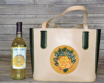 Könnte beinhalten: Eine beige Ledertasche mit grünem Lederbesatz und gelben Nähten. Die Tasche hat ein kreisförmiges Design mit einer gelben Rose und grünen Blättern auf der Vorderseite. Die Tasche steht neben einer Flasche Weißwein mit einem Etikett, auf dem "Dizzy Daisy Winery" steht.