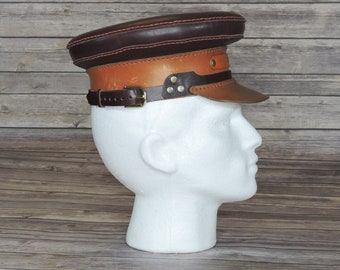 Könnte beinhalten: Eine braune Lederkappe mit einem hellbraunen Lederband und einer Messing-Schnalle. Die Kappe hat eine rote Nahtdetail um den Rand.