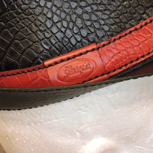 K&ouml;nnte beinhalten: Nahaufnahme eines schwarzen Lederriemens mit roten N&auml;hten und einem roten Lederflicken mit dem in Wei&szlig; gepr&auml;gten Text "Garcia's Leather Works".