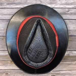K&ouml;nnte beinhalten: Ein schwarzer Lederhut im Fedora-Stil mit einem roten Lederband. Der Hut hat eine strukturierte, krokodilgepr&auml;gte Krone.