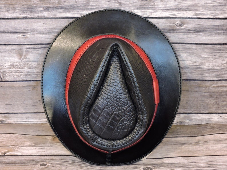 K&ouml;nnte beinhalten: Ein schwarzer Lederhut im Fedora-Stil mit einem roten Lederband. Der Hut hat eine strukturierte, krokodilgepr&auml;gte Krone.