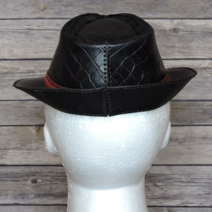 K&ouml;nnte beinhalten: Schwarzer Leder-Fedora-Hut mit rotem Lederband und einem gesteppten Muster auf der Krone. Der Hut wird von hinten gezeigt.