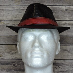 K&ouml;nnte beinhalten: Ein schwarzer Leder-Fedora-Hut mit einem roten Lederband. Der Hut hat eine Kroko-Pr&auml;gung.