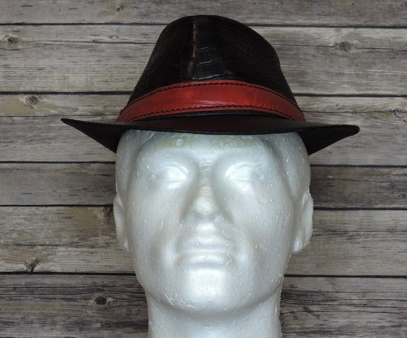 K&ouml;nnte beinhalten: Ein schwarzer Leder-Fedora-Hut mit einem roten Lederband. Der Hut hat eine Kroko-Pr&auml;gung.