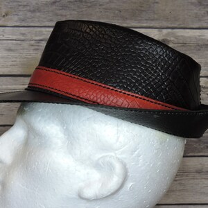 K&ouml;nnte beinhalten: Ein schwarzer Leder-Fedora-Hut mit einem roten Lederband. Der Hut hat eine strukturierte Oberfl&auml;che, die an Krokodilleder erinnert.