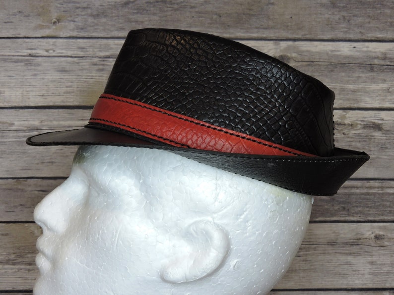 K&ouml;nnte beinhalten: Ein schwarzer Leder-Fedora-Hut mit einem roten Lederband. Der Hut hat eine strukturierte Oberfl&auml;che, die an Krokodilleder erinnert.