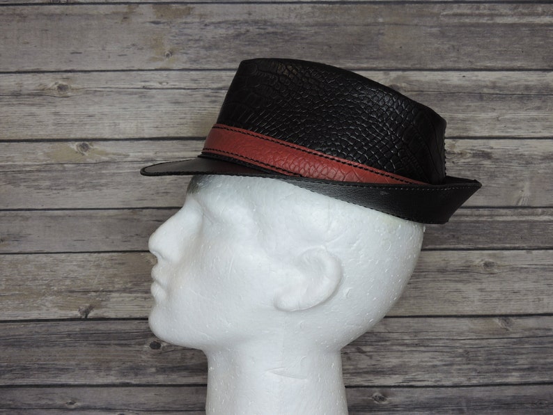 K&ouml;nnte beinhalten: Ein schwarzer Leder-Fedora-Hut mit einem roten Lederband um die Krone.