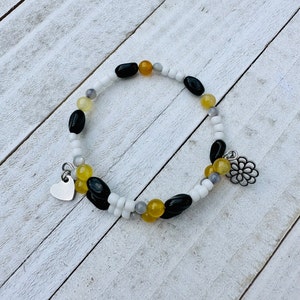 May include: A white, black and yellow beaded bracelet with a silver heart charm and a silver flower charm.