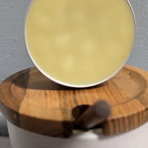 K&ouml;nnte beinhalten: Ein runder, blassgelber Balsam in einer silbernen Metalldose, der auf einem wei&szlig;en Keramik-Honigtopf mit Holzdeckel ruht, aus dessen Mitte ein kleiner Holzstiel ragt. Das Wort HONEY ist in schwarzen Gro&szlig;buchstaben auf den Topf gedruckt.