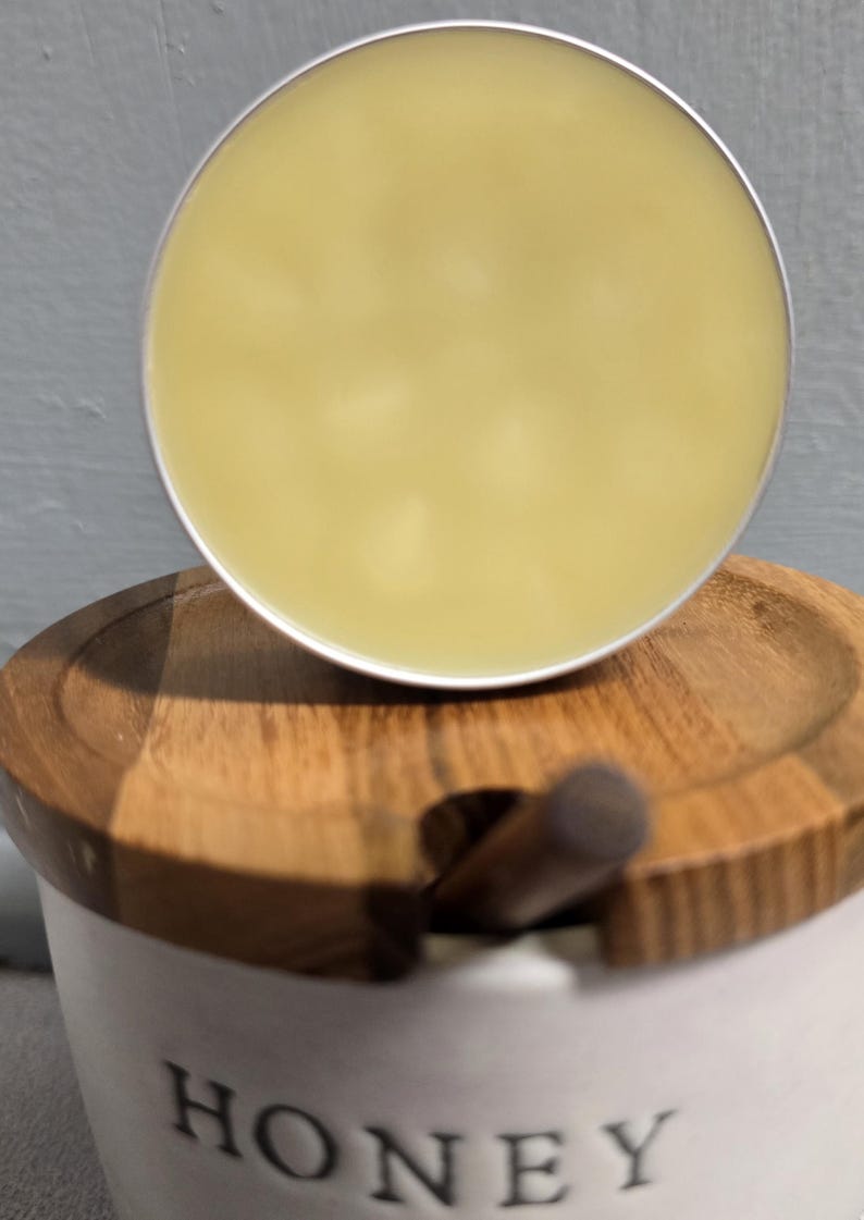 K&ouml;nnte beinhalten: Ein runder, blassgelber Balsam in einer silbernen Metalldose, der auf einem wei&szlig;en Keramik-Honigtopf mit Holzdeckel ruht, aus dessen Mitte ein kleiner Holzstiel ragt. Das Wort HONEY ist in schwarzen Gro&szlig;buchstaben auf den Topf gedruckt.