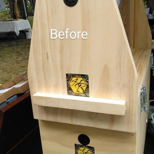 K&ouml;nnte beinhalten: Ein heller Bienenstockkasten aus Holz mit dem Wort Before in wei&szlig;er Schrift. Die Struktur umfasst zwei kreisf&ouml;rmige schwarze Bel&uuml;ftungs&ouml;ffnungen und zwei quadratische Aufkleber mit einem gelben Sechseck und den schwarzen Initialen HB.