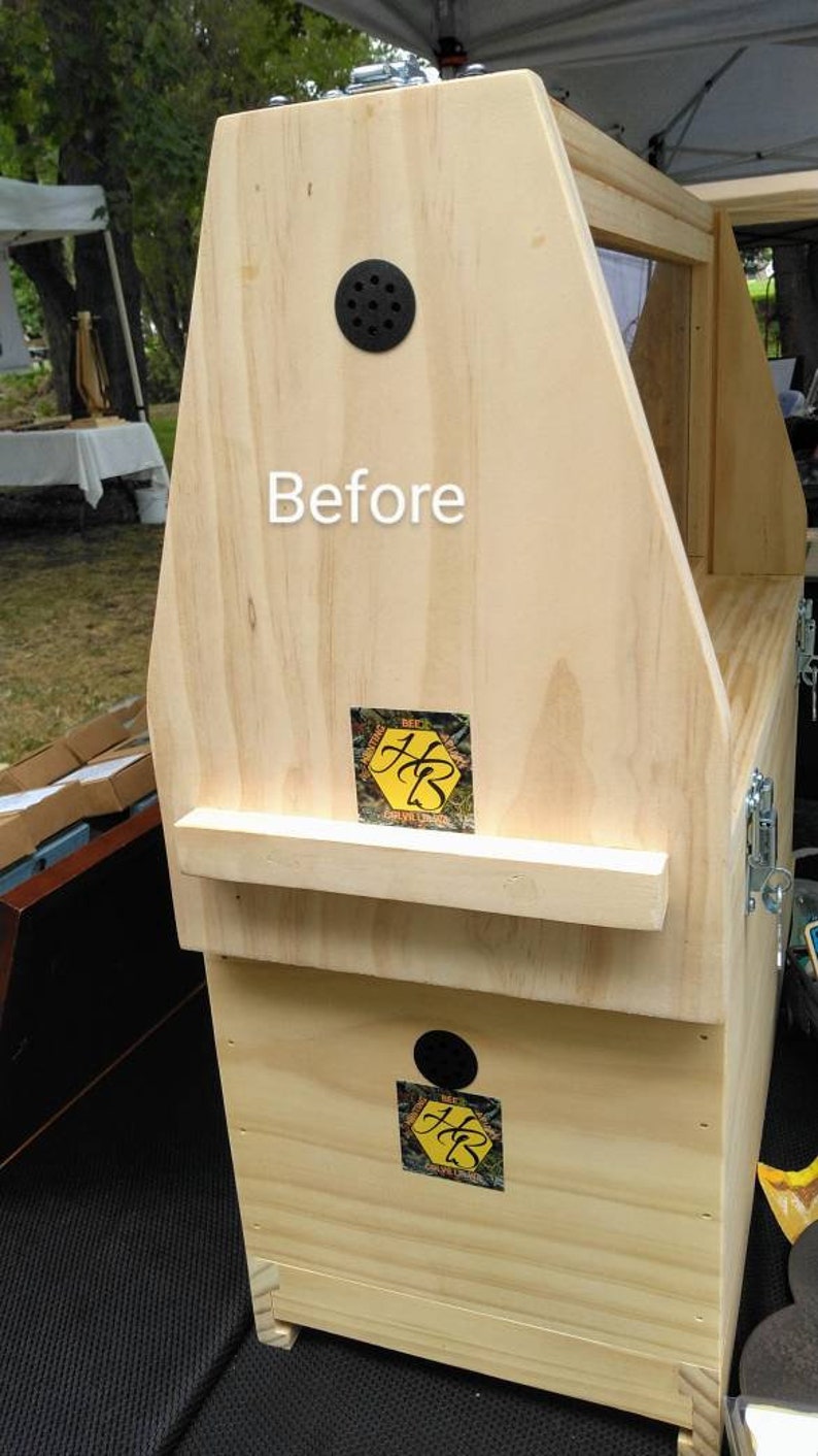 K&ouml;nnte beinhalten: Ein heller Bienenstockkasten aus Holz mit dem Wort Before in wei&szlig;er Schrift. Die Struktur umfasst zwei kreisf&ouml;rmige schwarze Bel&uuml;ftungs&ouml;ffnungen und zwei quadratische Aufkleber mit einem gelben Sechseck und den schwarzen Initialen HB.