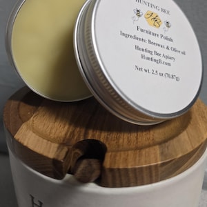 K&ouml;nnte beinhalten: Eine runde silberne Dose mit Hunting Bee M&ouml;belpolitur, wei&szlig;em Etikett mit Bienenlogo, Zutatenliste und einem Nettogewicht von 70,87g. Die Dose steht auf einem wei&szlig;en Keramik-Honigtopf mit Holzdeckel und einem kleinen h&ouml;lzernen Honigl&ouml;ffel.