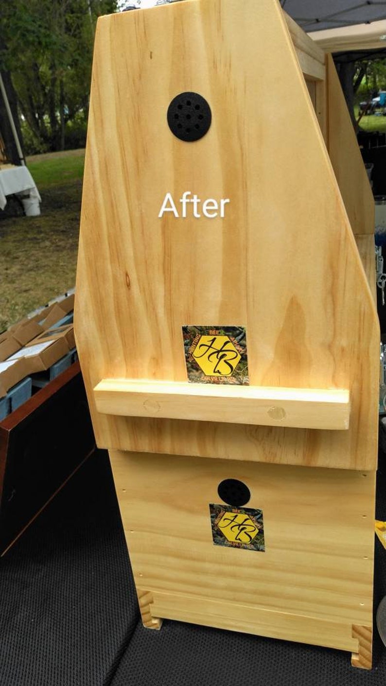 K&ouml;nnte beinhalten: Eine helle Bienenstockstruktur aus Holz mit dem in Wei&szlig; gedruckten Wort After. Sie enth&auml;lt zwei schwarze, kreisf&ouml;rmige Bel&uuml;ftungs&ouml;ffnungen und zwei quadratische Aufkleber mit gelbem Wabenmuster und den schwarzen Buchstaben HB auf der Vorderseite.