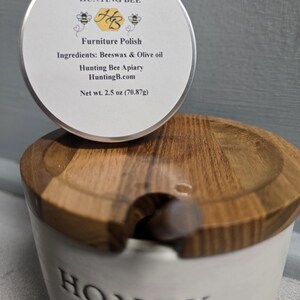 K&ouml;nnte beinhalten: Eine runde Metalldose mit Hunting Bee M&ouml;belpolitur, wei&szlig;es Etikett mit Bienen, Text Hunting Bee, Furniture Polish, Ingredients: Beeswax & Olive oil und Net wt. 2.5 oz (70,87g), platziert auf einem wei&szlig;en Keramik-Honigtopf mit einem Holzdeckel.
