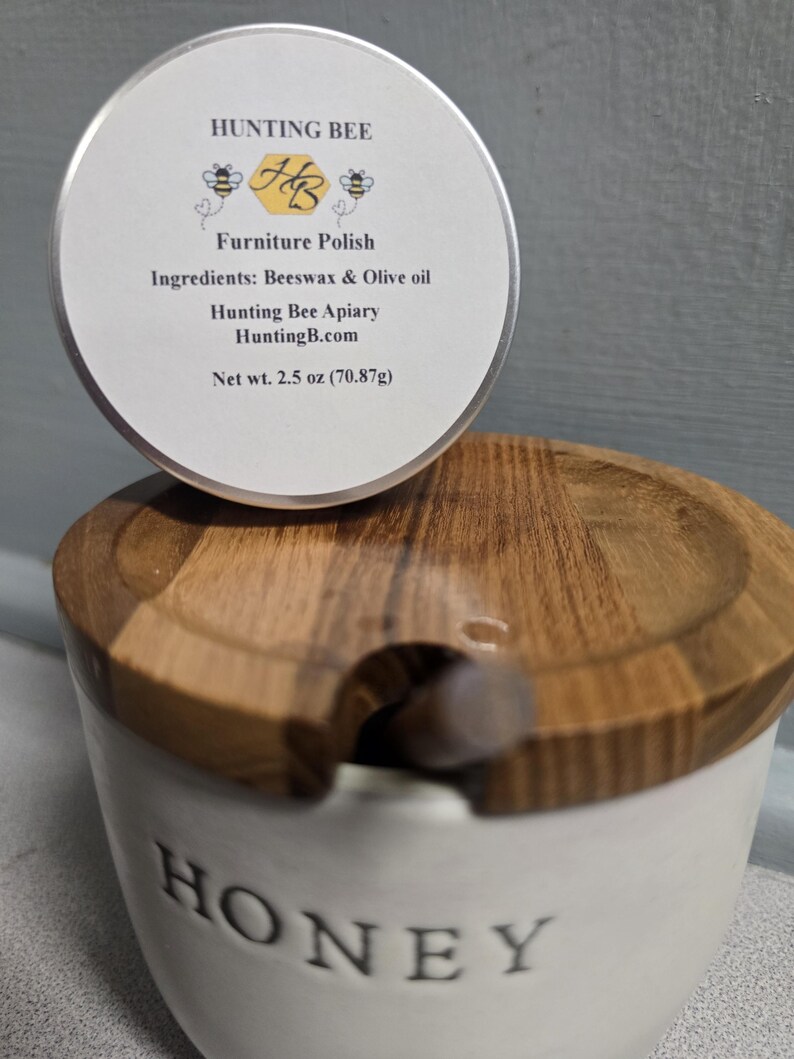 K&ouml;nnte beinhalten: Eine runde Metalldose mit Hunting Bee M&ouml;belpolitur, wei&szlig;es Etikett mit Bienen, Text Hunting Bee, Furniture Polish, Ingredients: Beeswax & Olive oil und Net wt. 2.5 oz (70,87g), platziert auf einem wei&szlig;en Keramik-Honigtopf mit einem Holzdeckel.