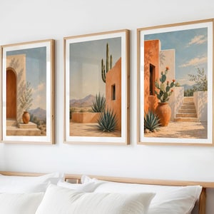 Può includere: Tre stampe artistiche incorniciate raffiguranti paesaggi desertici con edifici in terracotta, cactus e montagne sotto un cielo blu. Le stampe sono in cornici di legno chiaro e sono appese sopra un letto con biancheria da letto bianca.