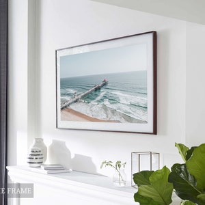 May include: A framed photograph of a pier extending into the ocean with waves crashing against the shore. The photo is in a brown frame and is hanging on a white wall. The photo is displayed above a white mantelpiece with a vase and books on it.