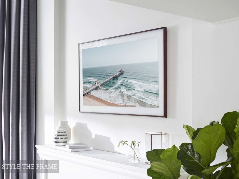 May include: A framed photograph of a pier extending into the ocean with waves crashing against the shore. The photo is in a brown frame and is hanging on a white wall. The photo is displayed above a white mantelpiece with a vase and books on it.