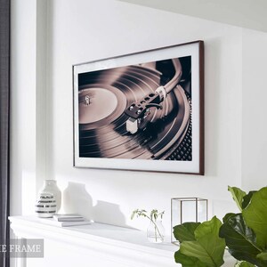 Peut inclure: Une photographie encadr&eacute;e d'un tourne-disque avec un disque sur le plateau. Le disque est noir et le plateau est argent&eacute;. La photo est dans un cadre brun et est accroch&eacute;e &agrave; un mur blanc.