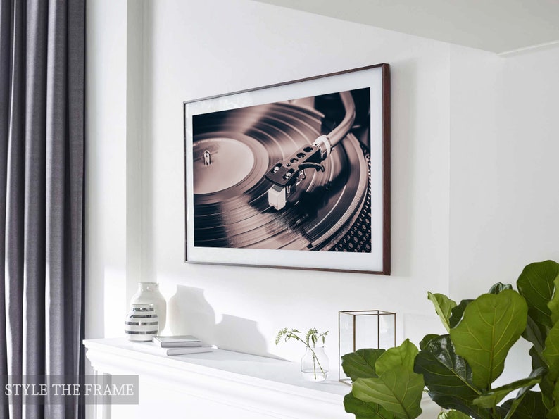Peut inclure: Une photographie encadr&eacute;e d'un tourne-disque avec un disque sur le plateau. Le disque est noir et le plateau est argent&eacute;. La photo est dans un cadre brun et est accroch&eacute;e &agrave; un mur blanc.