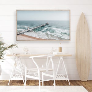 May include: A framed photograph of a pier extending out into the ocean with waves crashing against the shore. The photo is hanging on a white wall above a white desk with a white chair.