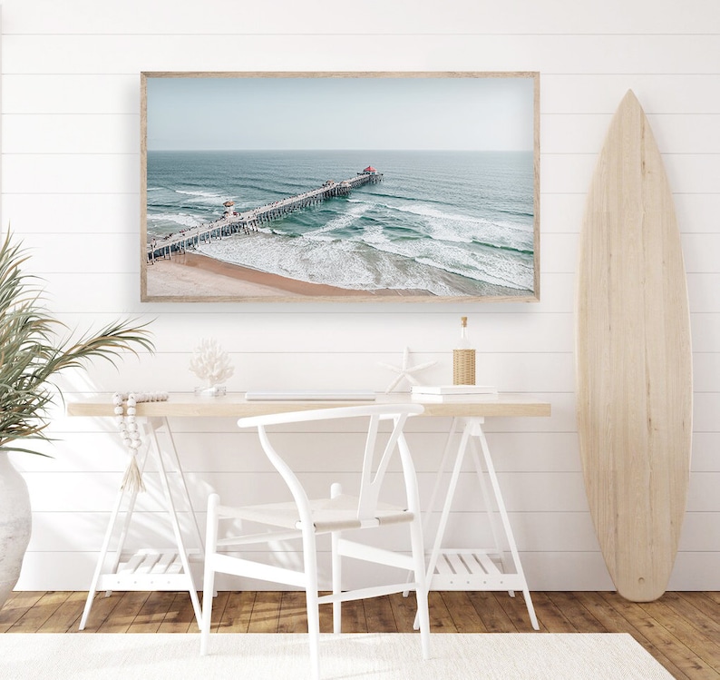 May include: A framed photograph of a pier extending out into the ocean with waves crashing against the shore. The photo is hanging on a white wall above a white desk with a white chair.