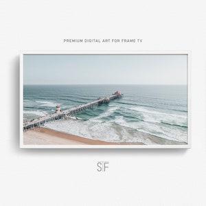 May include: An aerial view of a pier extending out into the ocean with white capped waves crashing on the sandy beach. The pier has a red roof at the end.