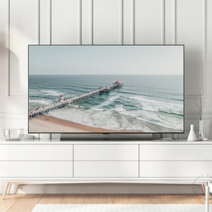 May include: A television displaying a photograph of a pier extending into the ocean with waves crashing against the shore. The pier is made of wood and has a red roof. The water is a light blue color and the sky is a light gray color.